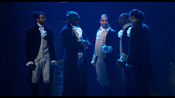 Five men in 18th-century period costumes standing together on a dark, blue-lit stage.