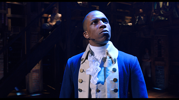 A man in a blue and white period costume looks upward on a dark stage.