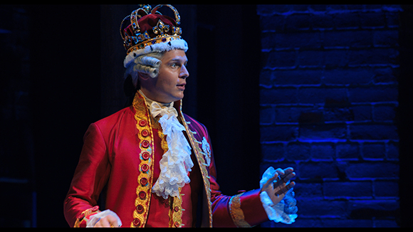 A man in a red royal coat and crown gestures with his hands against a dark blue brick background.