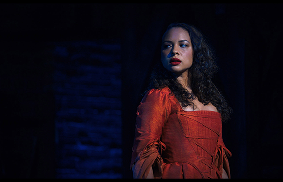 A woman with curly hair wearing a red corset-style dress looks into the distance against a dark blue background.
