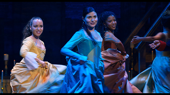 Three women in colorful 18th-century style gowns dancing under blue stage lighting.
