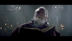 An elderly man with a white beard in a brown robe looks upward while holding an open book.
