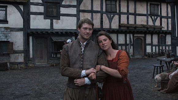 A man and woman in historical attire stand close together in a courtyard with Tudor-style buildings.
