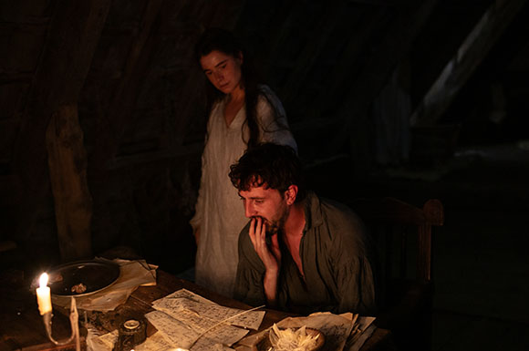 A man sits at a wooden table reading papers by candlelight while a woman stands behind him in a dark room.