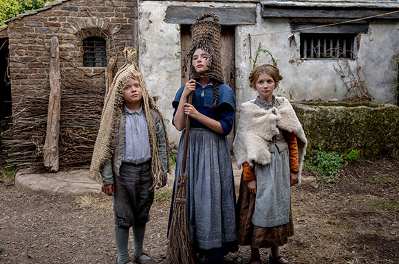 Three children in rustic clothing stand outside a stone cottage, wearing woven straw headpieces.