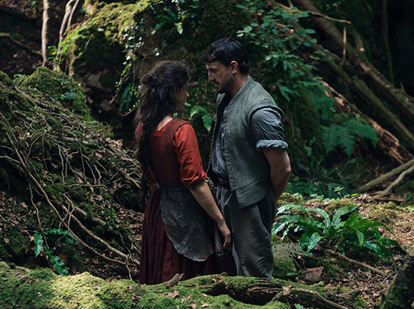 A man and a woman in rustic clothing stand facing each other in a dense, green forest.