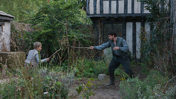 A man in period clothing points a stick at a boy holding a stick in a lush garden near a Tudor-style house.