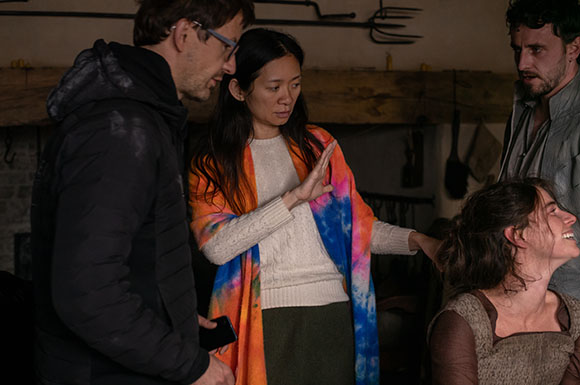 A woman in a white sweater and colorful shawl gestures toward a man while others look on in a dark room.