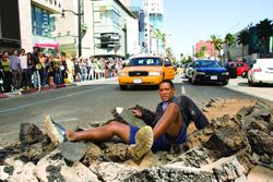 A man sits in a pile of rocks in the middle of a city street with cars and pedestrians in the background.