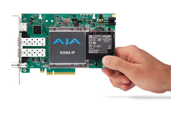A person's hand holds a green AJA KONA IP circuit board with various ports and components.