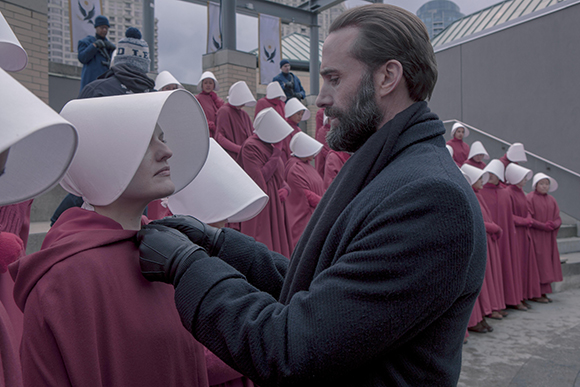 A man in a dark coat adjusts the collar of a woman wearing a red robe and a large white bonnet.