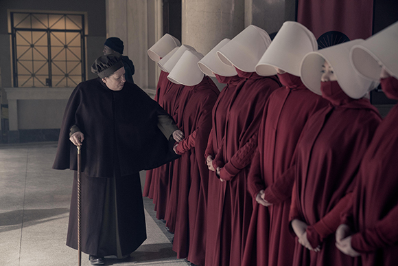 An older woman with a cane walks past a row of women wearing red robes and large white winged headpieces.