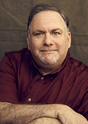 A middle-aged man with short brown hair and a grey goatee wearing a maroon button-down shirt.