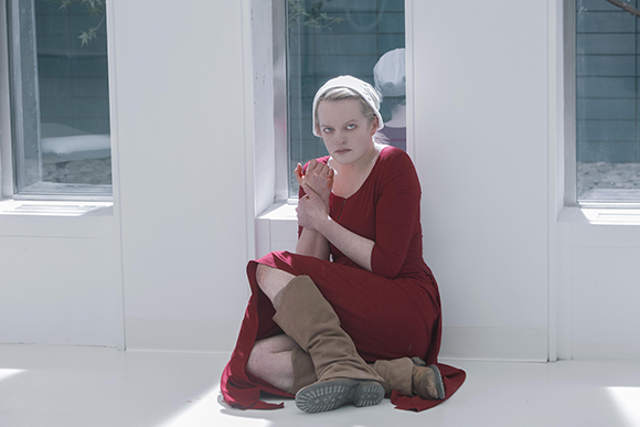 A woman in a long red dress and white bonnet sits on a white floor against a white wall with windows.