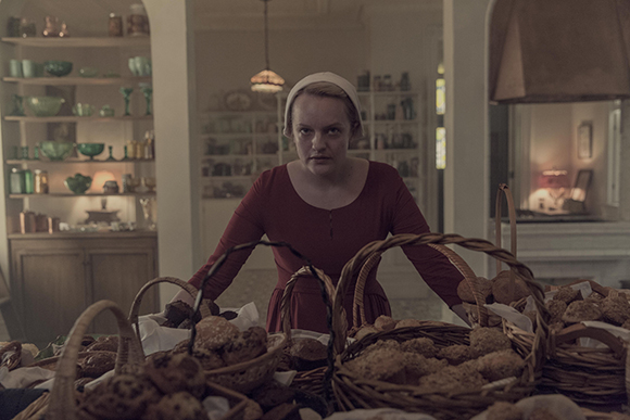 A woman in a red dress and white cap leans over several wicker baskets filled with various types of bread.