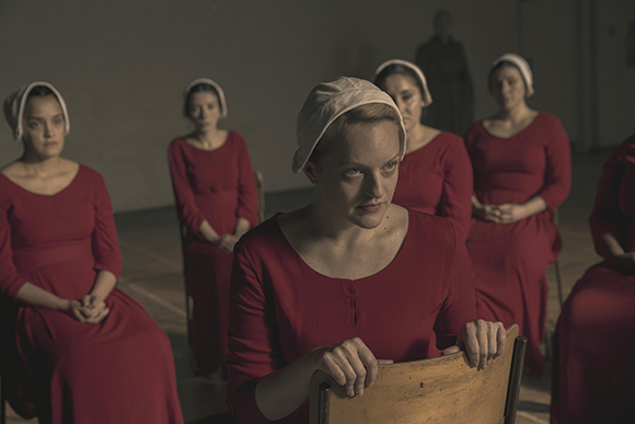 Several women wearing red dresses and white bonnets sit in a dimly lit room.