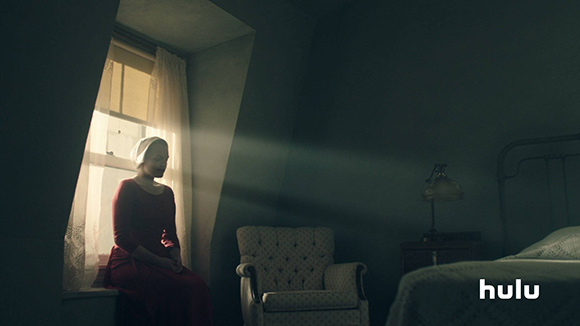 A woman in a red dress and white headpiece sits in a window nook as light streams into a dark room.