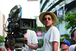 A man wearing a straw hat and sunglasses stands beside a large professional motion picture camera on a film set.