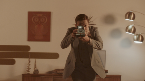 A man in a grey suit holds a vintage camera up to his eye, aiming it directly at the camera in a dimly lit room.