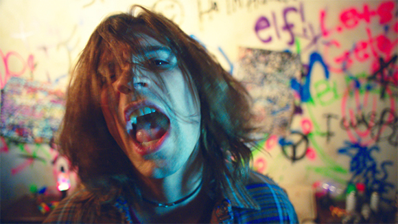 A person with long hair singing with an open mouth against a wall covered in colorful graffiti.