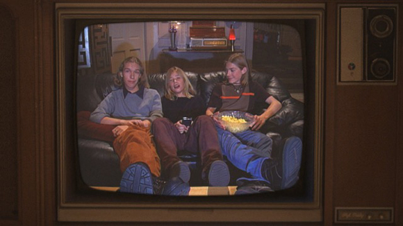 Three teenagers sit on a dark leather couch holding snacks while watching a screen.