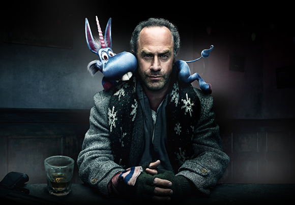 A man in a patterned sweater sits at a bar with a blue cartoon donkey perched on his shoulder.