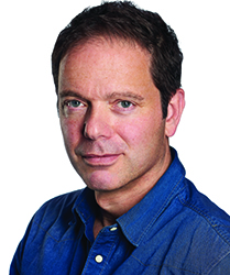 A man with short dark hair and light eyes wearing a blue denim button-down shirt against a white background.