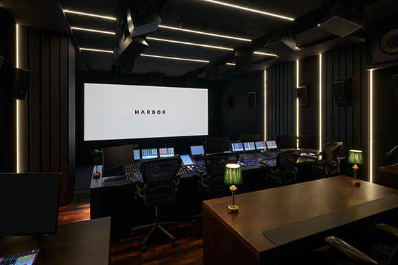 A dark professional sound mixing studio with multiple monitors, large consoles, and linear ceiling lighting.