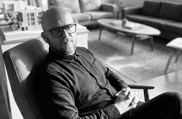 A bald man with glasses and a beard sits in a chair in a modern living room.