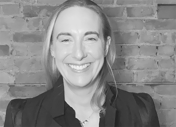 A smiling woman with long hair wearing a dark blazer in front of a brick wall.