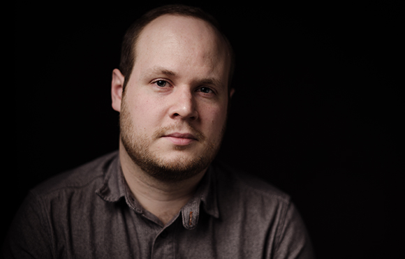 A man with short hair and a light beard looks directly at the camera against a dark background.