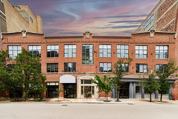 A wide red brick commercial building with large windows and a street-level entrance under a pink sunset sky.