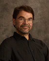 A man with dark hair, glasses, and a salt-and-pepper beard wearing a black button-down shirt.