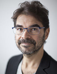 A man with dark wavy hair, glasses, and a salt-and-pepper beard looking toward the camera.