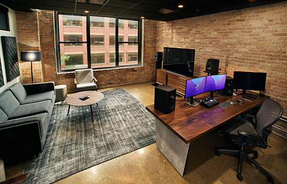 A home office with a large wooden desk, dual monitors, a sofa, and a coffee table in a brick-walled room.