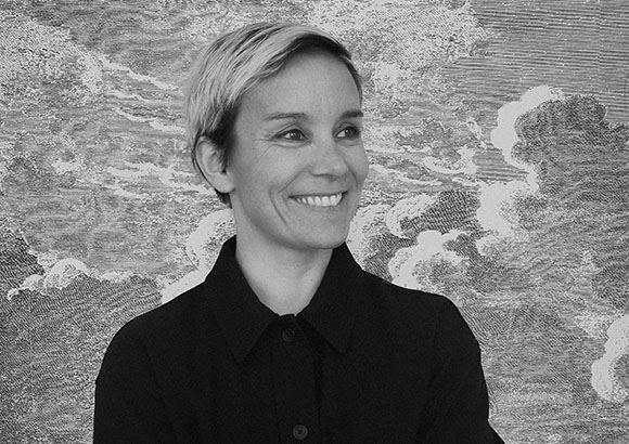 A smiling woman with short blonde hair wearing a dark button-down shirt against a textured backdrop.
