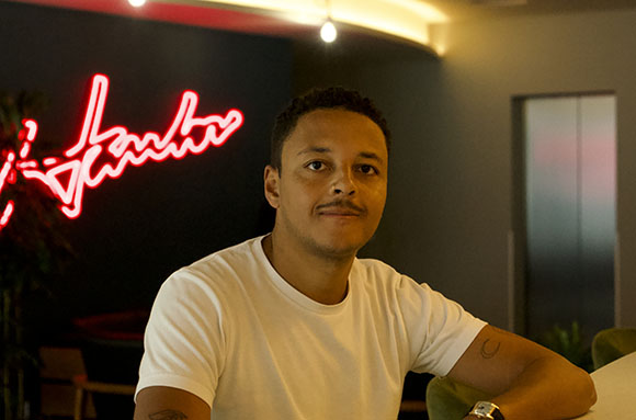 A man in a white t-shirt sits in a dimly lit room with a red neon sign in the background.