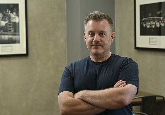 A man with short brown hair and a mustache stands with arms crossed in an office setting.