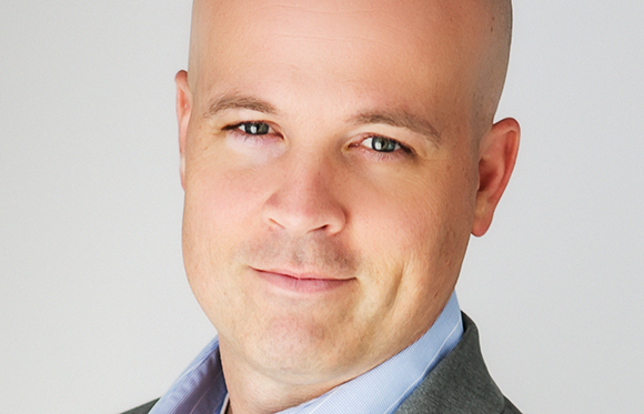 A close-up portrait of a bald man with light eyes wearing a light blue shirt and grey blazer.