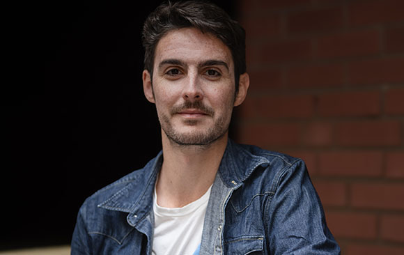 A man with dark hair and light stubble wearing a blue denim jacket over a white t-shirt.