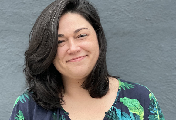 A smiling woman with dark hair wearing a dark blue tropical print shirt against a gray wall.