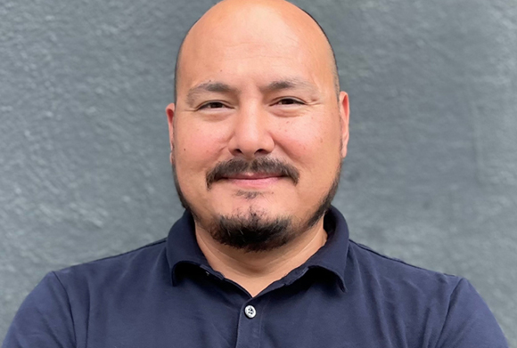 A bald man with a dark goatee and mustache wearing a navy blue button-down shirt.
