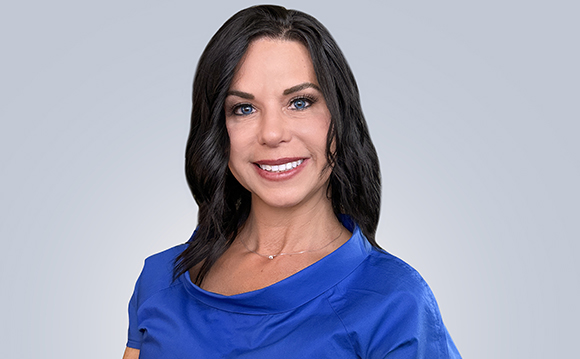 A woman with long dark hair and blue eyes smiling in a blue top against a gray background.