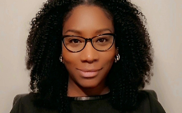 A woman with dark curly hair and black-rimmed glasses smiling slightly against a plain background.