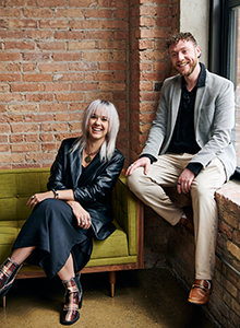 A woman in a black leather jacket sits on a green sofa while a man in a grey blazer sits on a windowsill.