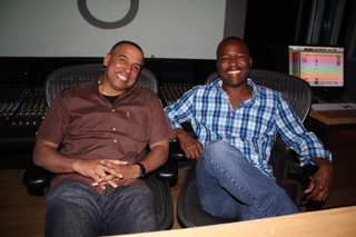 Two men sitting in chairs in a studio with a mixing console and monitor in the background.