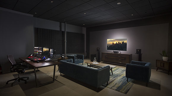 A dark control room with a desk featuring multiple monitors and a mixing console, facing a large screen on a console.
