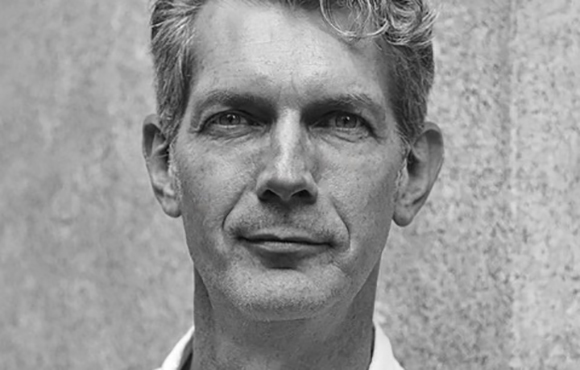 Black and white close-up portrait of a man with wavy hair looking directly at the camera.