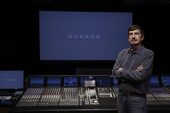 A man with arms crossed stands before a large audio mixing console in a dark studio.