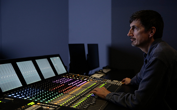 A man sits at a large, illuminated audio mixing console in a dimly lit studio.